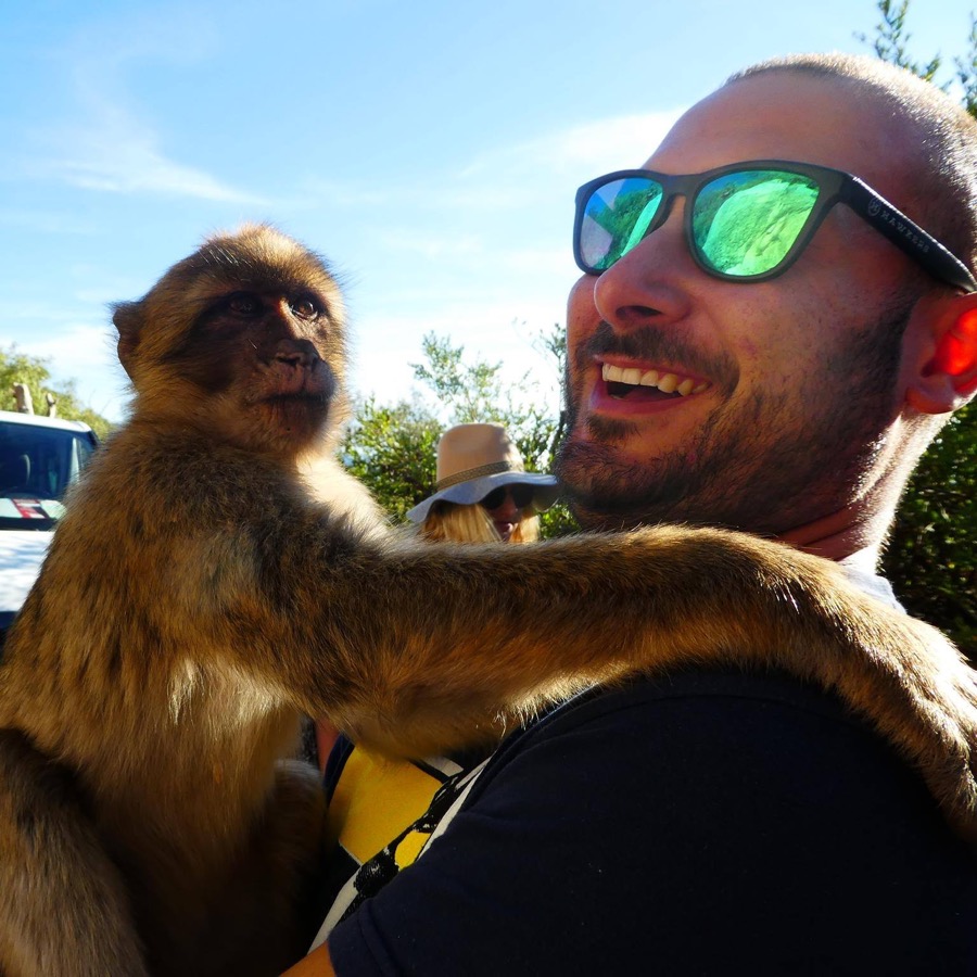 Jacopo Andrea Nuzzi smiling in mirrored sunglasses with a Barbary macaque sitting on his shoulder against a clear blue sky.